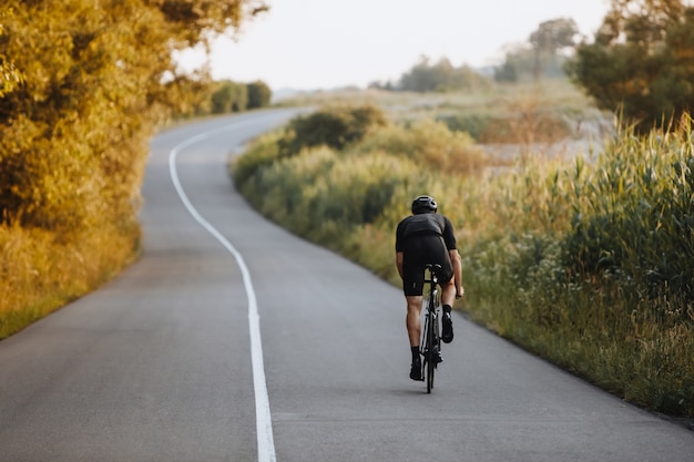 Back view of strong male cyclist with athletic body shape riding bike at the paved road among trees and green bushes