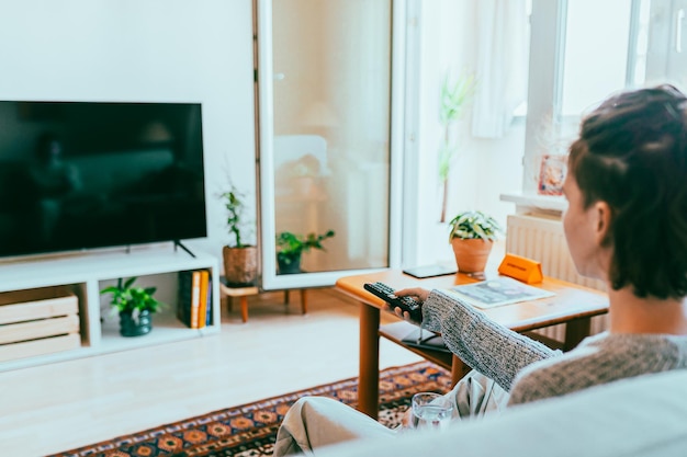 Back view of teenage girl sitting on sofa at home turning on TV with remote control