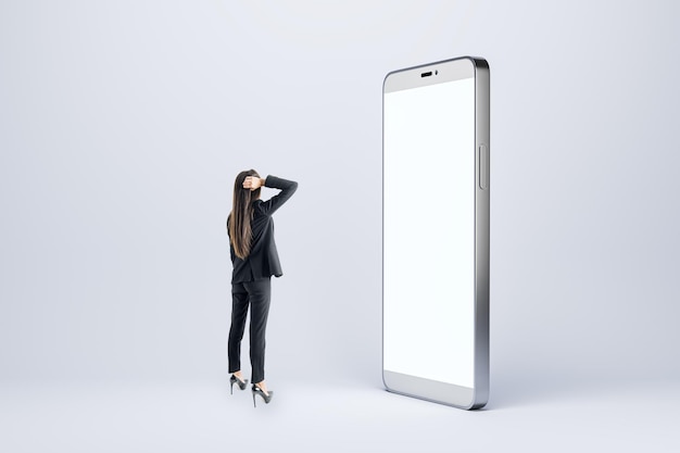 Photo back view of thoughtful young businesswoman looking at big empty white smartphone on light background technology innovation and communication concept mock up