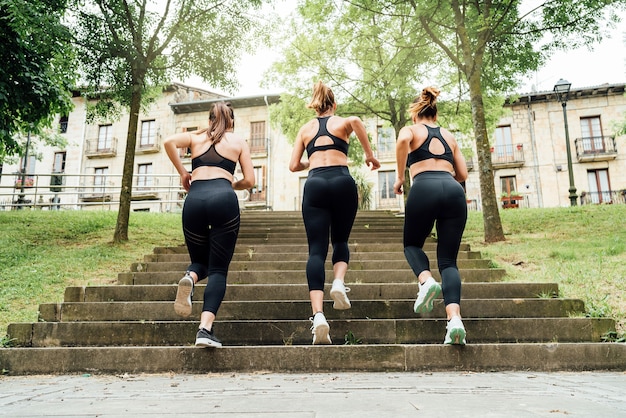 Vista posteriore tre belle donne che corrono su per le scale di un parco con molti alberi della città, tutte e tre vestite in abiti sportivi neri