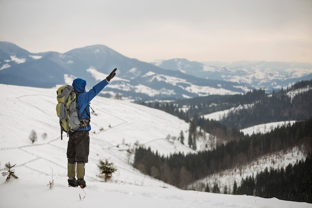 Back view of tourist hiker in warm clothing with backpack standing with raised arms on mountain clearing on copy space background of woody mountain ridge and cloudy sky.