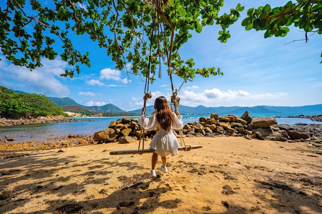 Back view of travel woman sitting on swing on the beach with palm trees and indian almond tree and turquoise sea on background Summer vacation tropical holidays Travel and lifestyle Concept