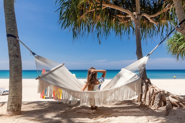 Back view of travel woman sitting on white hammock on sandy beach