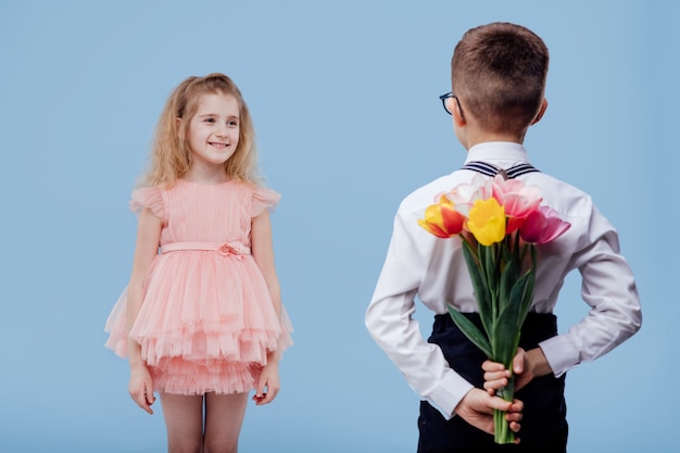 Back view, two little children boy with flowers and girl in pink dress, isolated on blue wall