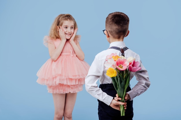 Вид сзади двух маленьких детей, мальчика с цветами и удивленной девочки в розовом платье, изолированных на голубом фоне...