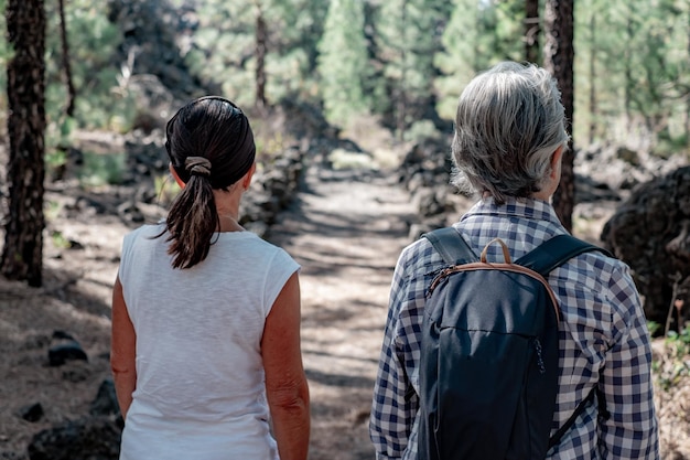 Back view of two mature women enjoying hike in the woods\
walking in footpath elderly retired females and adventure ageless\
concept