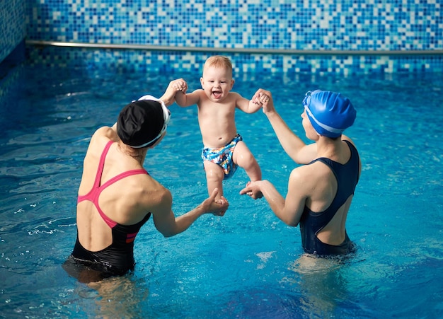 Back view of two women holding baby's hands and feet rising up laughing little boy from water after swimming in pool