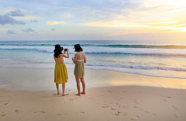 Вид сзади двух женщин сфотографировали красивые пейзажи морского заката с помощью мобильного телефона