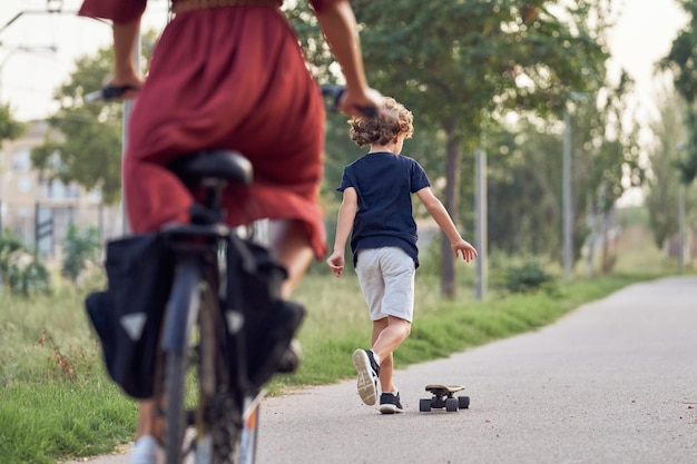 Back view of unrecognizable boy in casual clothes riding skateboard on asphalt road in park behind crop woman on bicycle