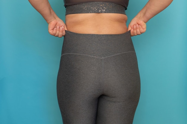 Back view of unrecognizable fat overweight woman standing in grey sports bra pulling on leggings showing buttocks