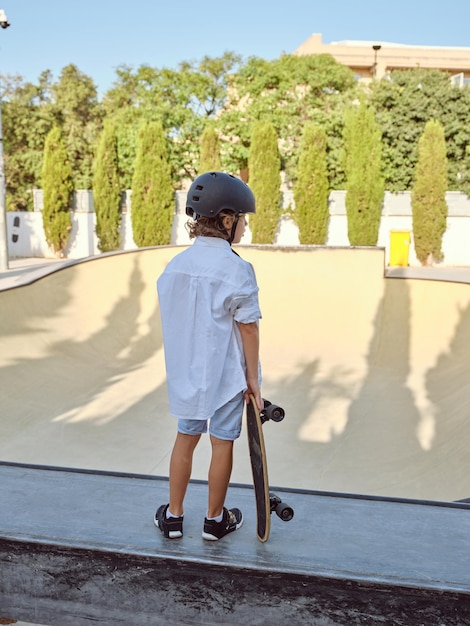 Back view of unrecognizable kid in casual clothes and helmet standing on ramp with skateboard in hand while looking at skate park at sunset