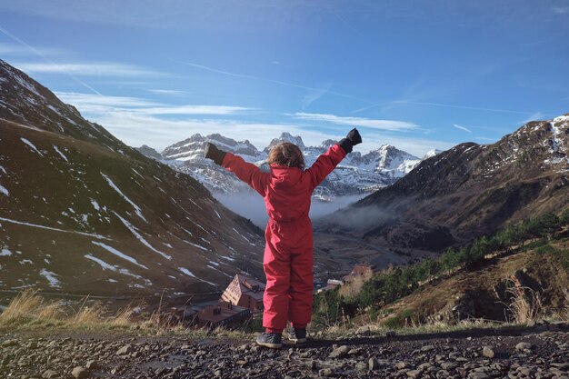 Back view of unrecognizable kid in red outerwear raising arms and enjoying freedom while standing near valley with village and admiring snowy mountains and cloudy blue sky