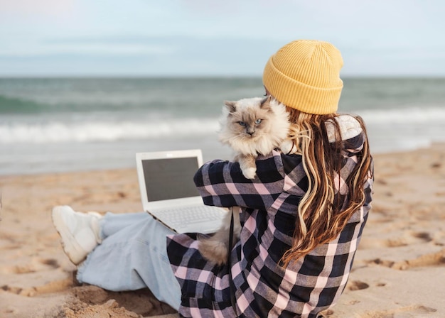 Back view of unrecognizable nomad digital woman in winter or autumn sitting on the beach using laptop with her cat on the shoulder