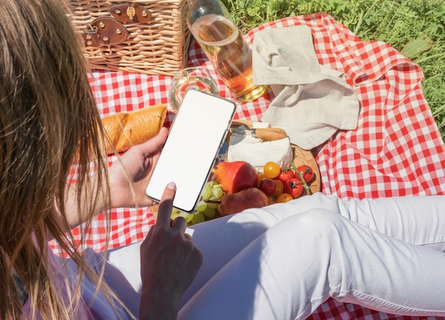 Back view of unrecognizable woman in white pants outside having picnic and using smartphone taking photo