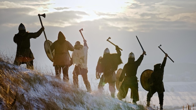 Back view. Vikings raise their weapons. The Vikings are dressed in chains, with swords, shields and helmets on their heads in a winter meadow.