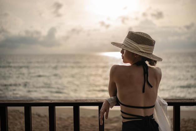 Back view of woman in black dress and straw hat standing on a balcony ocean view