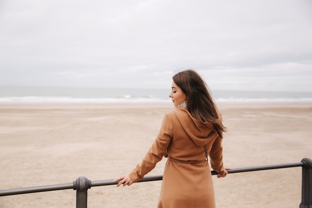 Back view of woman in brown coat standing along north sea and looking at the waves