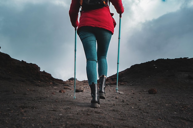 Back view of a woman hiker walking on the lava stone of the Volcano Etna in Sicily with trekking poles red vest and backpack wanderlust lifestyle concept closeup on the shoesxA