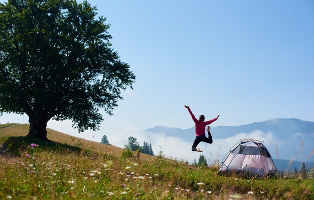 Punto di vista posteriore della donna che salta sulla collina di fioritura vicino alla tenda turistica e al grande albero sotto il chiaro cielo blu sulla mattina luminosa di estate sul fondo nebbioso delle montagne