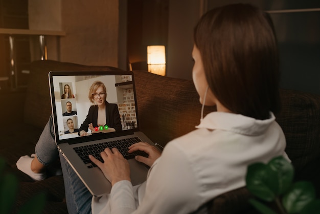 Back view of a woman lying at home on a sofa talking with her boss and other colleagues in a video call on a laptop. Businesswoman talks with coworkers on a webcam conference. A team having a meeting.