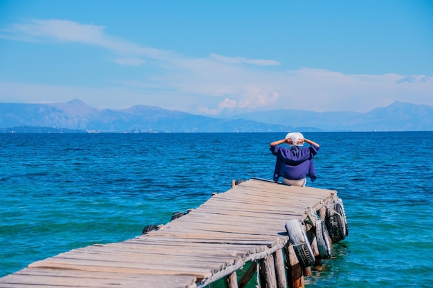 Back view of a woman sitting on a tiny wooden pier by the sea Young beautiful girl sitting on jetty Back view Amazing view on clear azure water and mountains on background Copy space