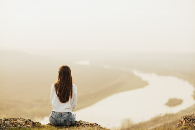丘の上に座って夕焼けの川を見ている女性の背面図。