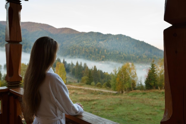 Back view of woman in spa bathrobe standing at wooden balcony of hotel resort room and enjoying