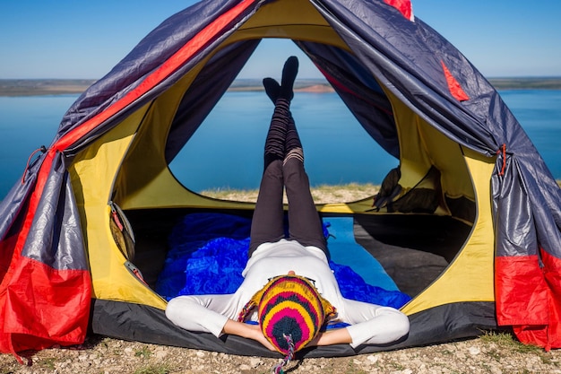 Back view of woman tourist in a funny hat from Nepal sitting in blue sleeping bag in a tent sunny good morning in the mountains lake