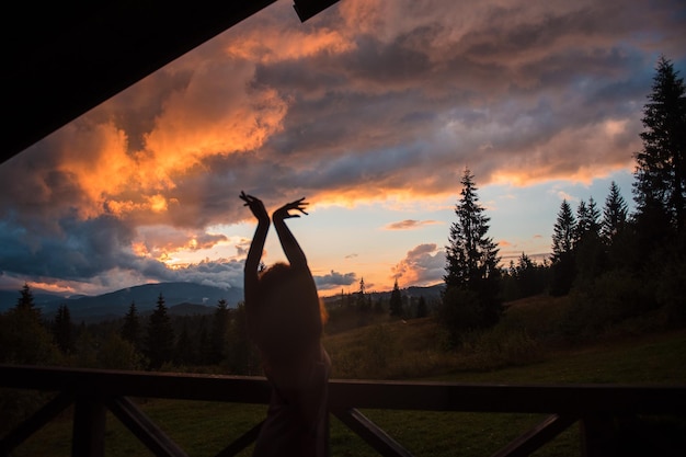 Back view of woman with open arms standing on open terrace and looking on beautiful sunset over majestic mountains Enjoyment from wild nature