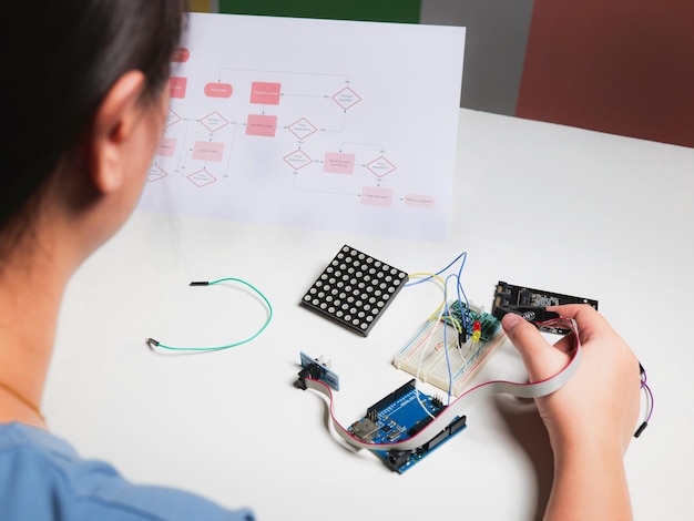 Back view young asian woman sitting at table using onboard programming debug improve binary algorithm code in workstation focusing on device and blurring people