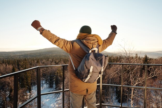 Back view of young backpacker keeping his arms raised