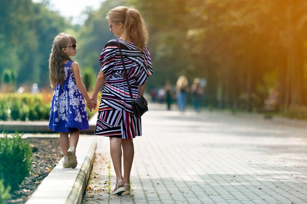 Back view of young blond long-haired slim attractive woman and small child girl in sunglasses and fashionable dresses walking holding hands by sunny summer park alley. Happy family relations concept.