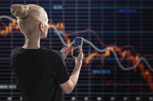 Back view of young blonde woman using mobile phone with falling red crisis forex chart on dark background Recession trade and downfall concept