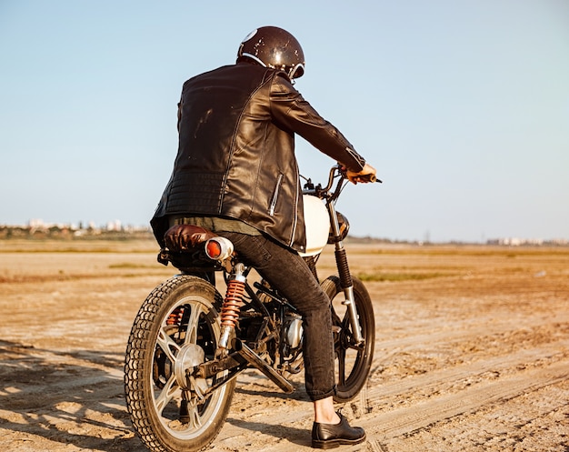 Back view of young brutal man in a black jacket and glasses driving retro motorcycle