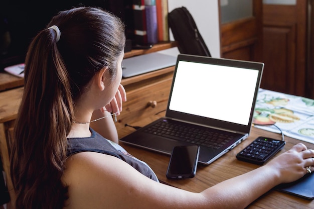 Back view of young caucasian woman at home looking at her laptop screen with one hand on the mouse