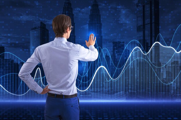 Back view of young european business man using abstract glowing\
blue business graph on blurry night city background financial\
growth market and stock concept double exposure