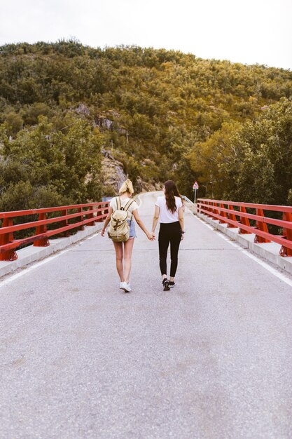 Back view of young girlfriends with backpack walking on the road over a bridge near the mountain.