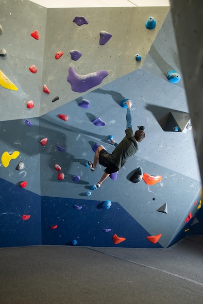 人工壁にぶら下がっている若い男の背面図。人工岩を保持し、壁を登ろうとしているジムでボルダリングをしているスポーツウェアのスポーティなアスリート。エクストリームスポーツと健康的なライフスタイルのコンセプト