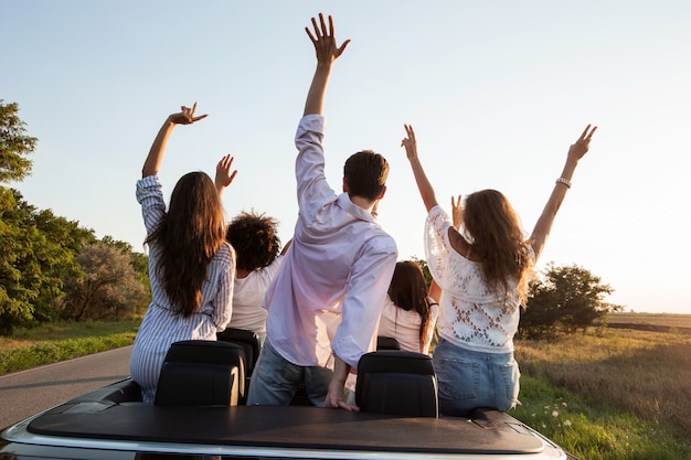 Back view. Young guys are sitting and holding hands up in a black cabriolet on the country road on a sunny day. .