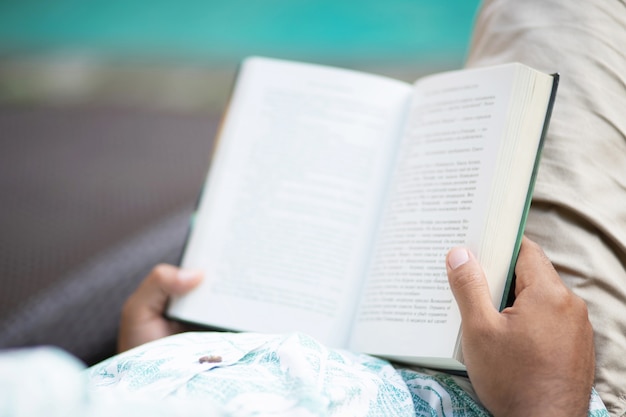 Back view of young man in casual clothes opening and reading a book and relaxing while lying on couch at by the swimming pool.