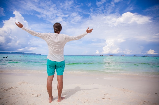 Back view of young man spread his arms standing on white sandy beach