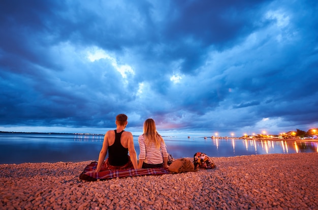 Back view of young man and woman sitting on rug on resort town pebble beach at dusk, enjoying view of cruise boats floating in calm blue water and dramatic cloudy evening sky