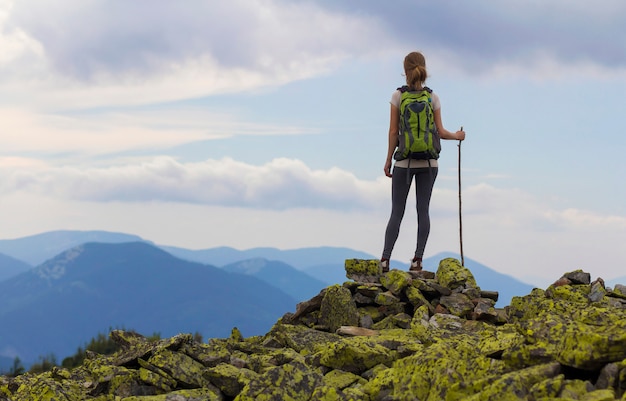 Punto di vista posteriore della ragazza turistica di giovane viaggiatore con zaino e sacco a pelo esile con il bastone che sta sulla cima rocciosa