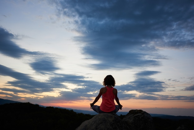 Back view of young slim woman sitting on big rock in yoga lotus pose on green tree tops dramatic evening sky at sunset background. Tourism, meditation and healthy lifestyle concept