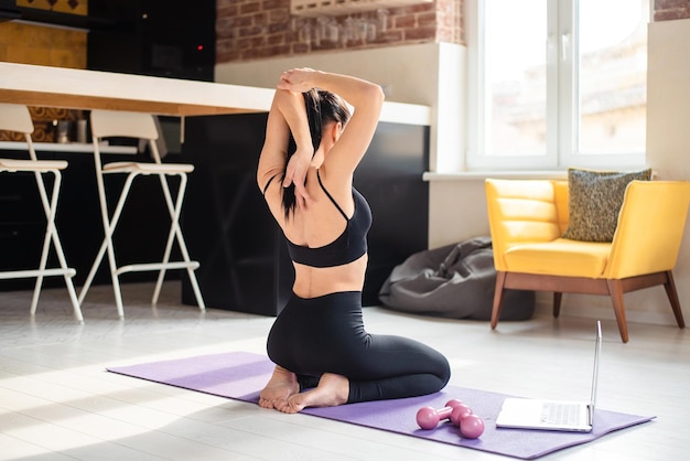 Back view of young sporty woman stretching arms and yoga mat Active brunette sitting on floor and using wireless laptop during training