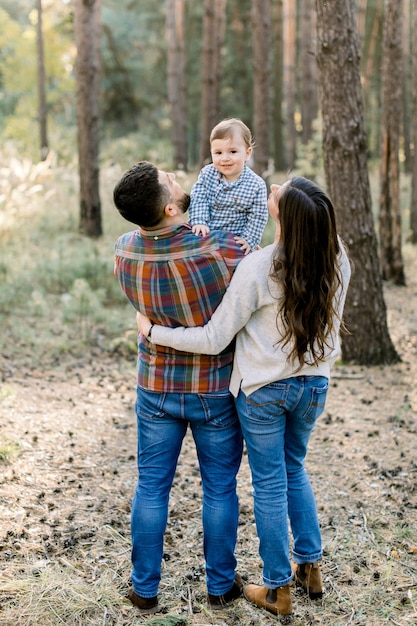 若いスタイリッシュな両親、父と母、彼らの小さな男の子を抱いて、カメラに微笑んでいる背面図。屋外の家族の肖像画