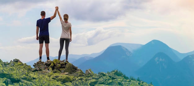 Back view of young tourist couple, athletic man and slim girl standing with raised arms holding hands on rocky mountain.