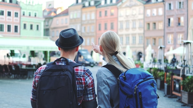 Back view of young tourists couple with bags checking map on central city square. They discussing their new destination.