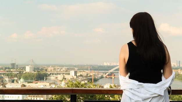 黒のTシャツと白のシャツを着た若い女性の背面図、ブルネットの旅行者は、観光客のための展望台のある高い丘から夏の晴れた日の街並みを眺めます。ビンテージ。