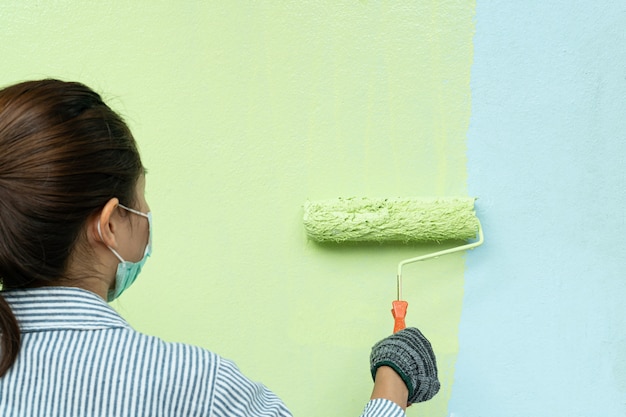 Punto di vista posteriore del pittore della giovane donna in camicia e guanti che dipingono una parete con il rullo di pittura.